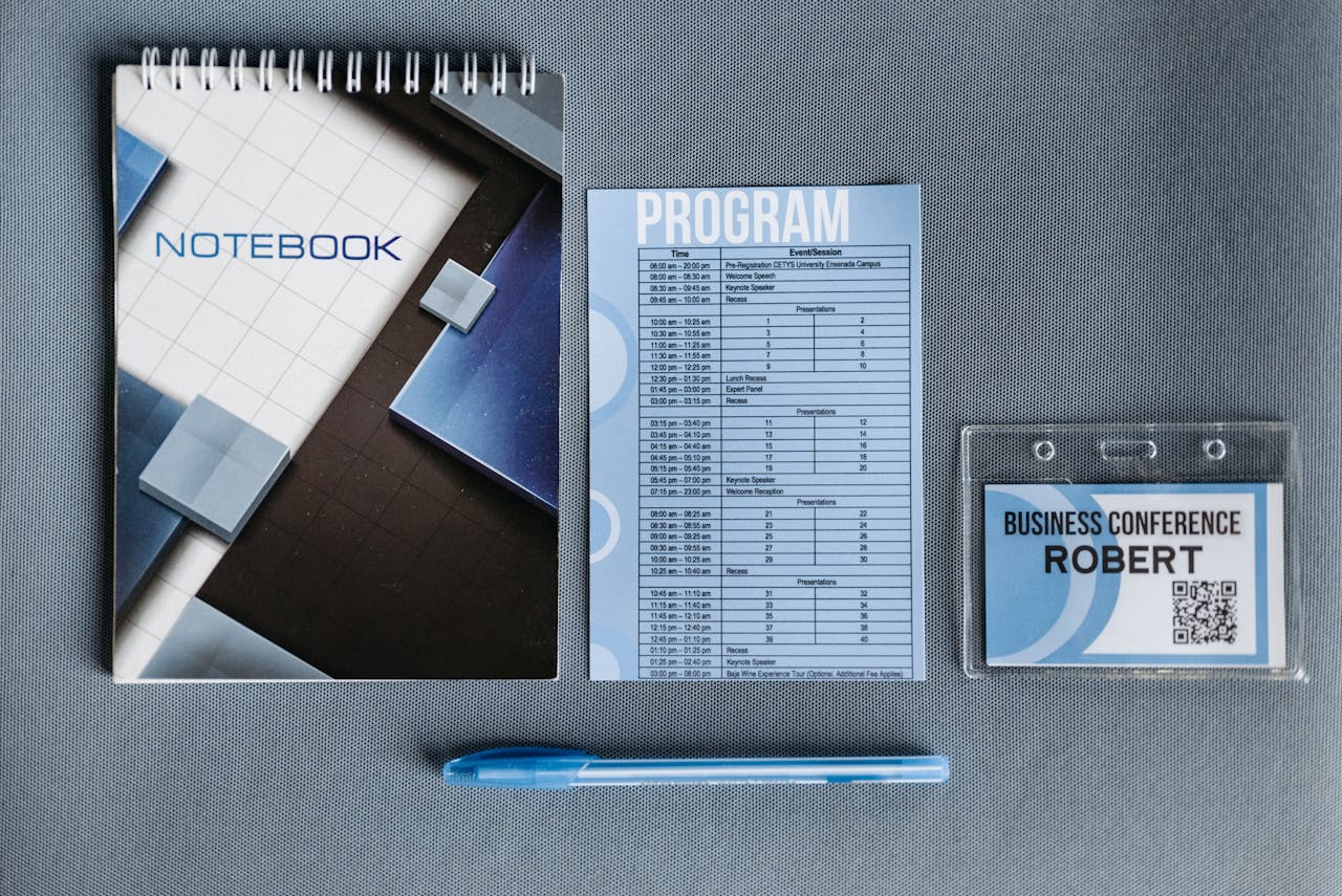 Flatlay of a notebook, conference badge, pen, and program on gray surface.